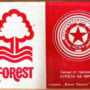 Puede incluir: A vintage matchbook with a red and white design. The left side shows a red tree logo and the word "FOREST". The right side features a red star inside a circular emblem, with Cyrillic text. A collectible item, possibly from a sporting event.
