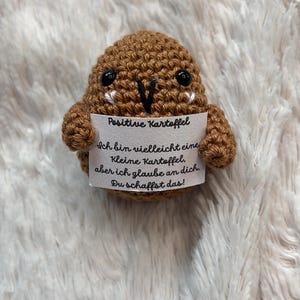 May include: A brown crocheted potato-shaped figure with black eyes and a white sign. The sign reads "Positive Kartoffel" and a German message of encouragement. The figure is on a fluffy white surface.