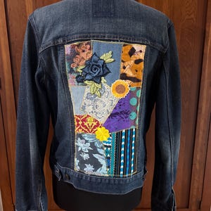 May include: A dark wash denim jacket with a patchwork design on the back. The patchwork features floral and geometric patterns in various colours, including blue, yellow, orange, and purple. The jacket has a classic collar, button closures, and long sleeves.