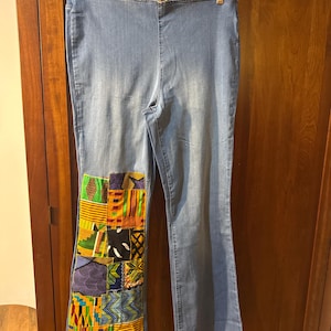 May include: Light blue denim pants with a patchwork design on the left leg. The patchwork features a variety of colorful African fabric patterns in shades of yellow, green, red, and blue. The pants are hanging on a clear hanger.