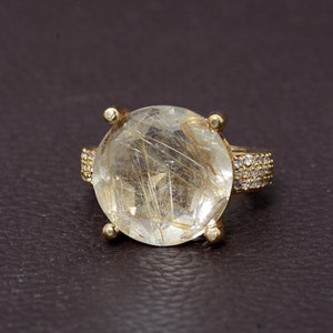 以下が含まれることがあります： 金色のリング。中央に金色のインクルージョンが入った、大きく丸い透明な宝石がセットされています。リングのバンドも金色で、小さな透明な宝石がちりばめられています。濃い茶色の背景。