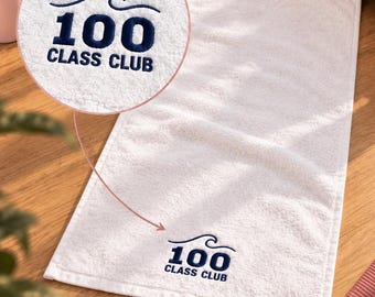 May include: A rectangular white towel featuring an embroidered dark blue logo that displays a stylized wave graphic above the text 100 CLASS CLUB. The embroidery is positioned near the bottom edge of the towel, with a circular inset showing detail.