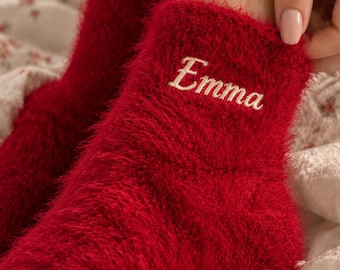 May include: A pair of vibrant red fuzzy ankle socks featuring a soft, textured material. The word Emma is embroidered in white cursive script on the outer side of the ankle area of the sock, providing a clear and legible personalized detail.
