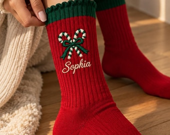 May include: Red ribbed crew socks featuring a dark green ruffled cuff, an embroidered design of two crossed candy canes tied with a green bow, and the name Sophia stitched in white cursive script on the front of the ankle area.