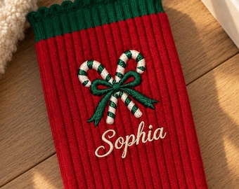 May include: A red ribbed sock with a green scalloped cuff. The sock features an embroidered design of two crossed candy canes tied with a green bow, positioned above the name Sophia embroidered in white cursive script lettering.