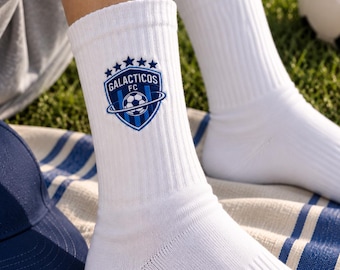 May include: White ribbed crew socks featuring an embroidered blue shield logo with the text Galacticos FC, five stars above the shield, and a ring encircling a soccer ball graphic in the center of the emblem, positioned on the upper ankle area.