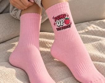May include: A pair of light pink ribbed crew socks featuring a graphic on the outer side of the left sock. The graphic includes the text PUMP UP THE PEACH in a bold, stylized font with a small illustration of a peach fruit and four-pointed stars.