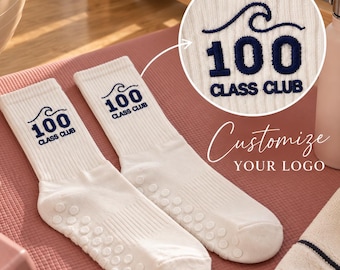 May include: A pair of white crew socks featuring a dark blue embroidered wave graphic above the text 100 CLASS CLUB. The soles of the socks have small circular white silicone grips for traction. A circular inset shows a close-up of the embroidery.