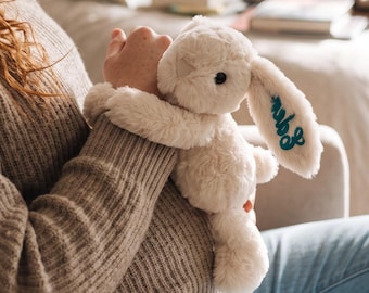 May include: A cream-colored plush rabbit toy with long ears, featuring dark teal cursive embroidery on one ear. The stuffed animal has a soft, textured fabric surface, black bead eyes, and a small stitched nose, designed as a cuddly companion.