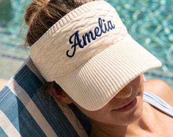 May include: A cream-colored knit visor featuring a textured cable-knit pattern on the headband and a ribbed brim. The name Amelia is embroidered in dark blue cursive script across the front center of the headband, providing a clear contrast.