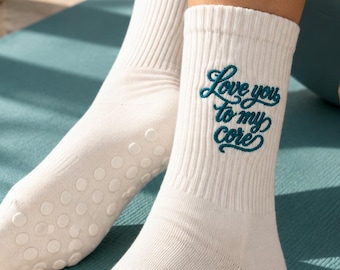 May include: A pair of white crew socks featuring ribbed cuffs and teal embroidered text that reads "Love you to my core." The soles of the socks are equipped with small, circular white silicone grip dots for traction.