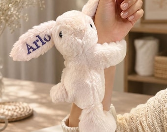 May include: A soft, cream-colored plush rabbit toy with long, floppy ears. One ear features the name Arlo embroidered in dark blue thread. The stuffed animal has a small, rounded tail, a stitched nose, and dark eyes, with a fuzzy, textured surface.