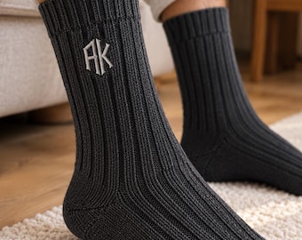 May include: A pair of dark charcoal gray ribbed knit socks featuring a white embroidered monogram with the letters AK on the outer side of the ankle. The socks have a thick, textured vertical ribbing pattern that extends from the cuff to the toe.