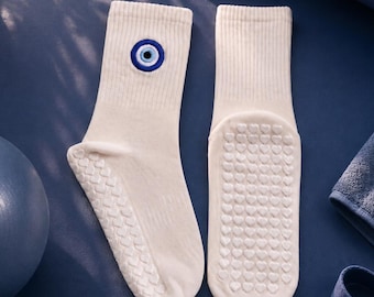 May include: A pair of cream-colored crew socks featuring a blue and white evil eye symbol embroidered on the ribbed cuff. The soles of the socks are covered in a repeating pattern of small, raised heart-shaped silicone grips for traction.