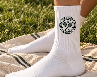 May include: White crew socks featuring a circular dark green emblem on the side. The emblem displays two crossed tennis rackets, a tennis ball, and the text Palm Valley Tennis Club enclosed within a laurel wreath border design.