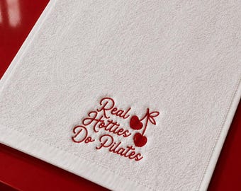 May include: A rectangular white towel featuring red embroidered text that reads Real Hotties Do Pilates, accompanied by a small red cherry graphic. The towel has a textured fabric surface and a finished border, presented in a clean, simple design.