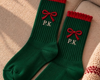 May include: A pair of dark green ribbed socks featuring a red ruffled cuff at the top. Each sock is embroidered with a red bow graphic and the letters P.K in white thread. The socks have a classic crew length design with a solid green foot.