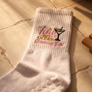 May include: A white crew sock featuring an embroidered martini glass graphic and the text "A Tiny Bit Older Birthday Girl" in pink and gold thread. The sock has a ribbed cuff and small white heart-shaped silicone grips on the sole for traction.