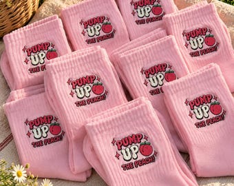 May include: A collection of light pink crew socks featuring an embroidered graphic of a peach and the text PUMP UP THE PEACH! in dark pink lettering. Each sock displays the same design on the upper cuff area, arranged in a group.