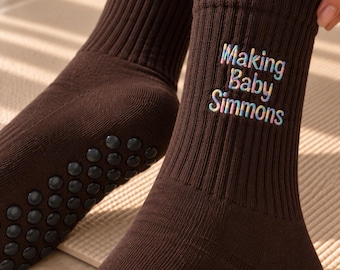 May include: A pair of dark brown ribbed crew socks featuring the text "Making Baby Simmons" embroidered in a multi-colored tie-dye style font. The soles of the socks are equipped with small, circular black non-slip silicone grips.