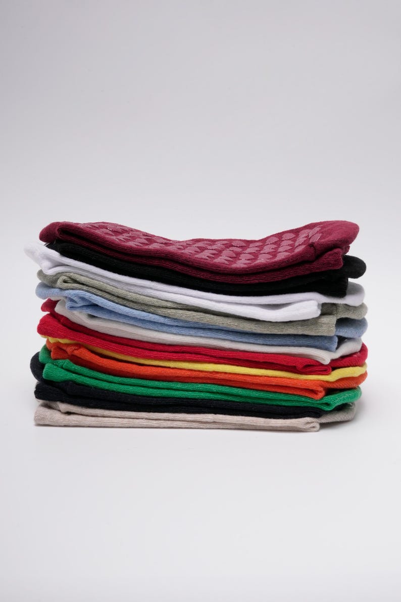 May include: A stack of folded socks in various solid colours including burgundy, black, white, grey, light blue, red, orange, yellow, green, and beige. The top burgundy pair features a textured pattern of small, raised grip dots on the fabric.