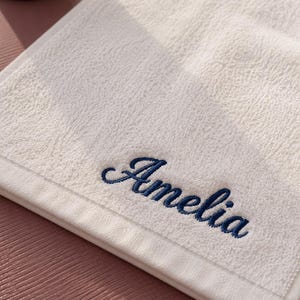May include: A white terry cloth towel featuring the name Amelia embroidered in dark blue cursive script. The towel has a textured surface and a smooth border along the edge, showcasing a clean and simple design for everyday bathroom use.