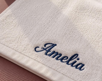 May include: A white terry cloth towel featuring the name Amelia embroidered in dark blue cursive script. The towel has a textured surface and a smooth border along the edge, showcasing a clean and simple design for everyday bathroom use.