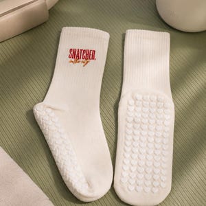 May include: A pair of cream-coloured ribbed crew socks featuring the red and orange text "SNATCHED in the city" on the upper ankle. The soles of the socks are covered in a white silicone grip pattern consisting of small, repeating heart shapes.