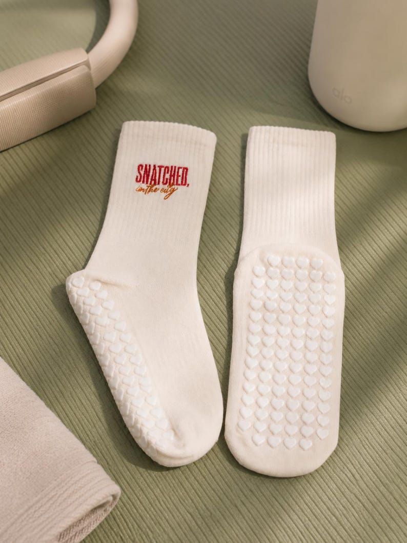 May include: A pair of cream-coloured ribbed crew socks featuring the red and orange text "SNATCHED in the city" on the upper ankle. The soles of the socks are covered in a white silicone grip pattern consisting of small, repeating heart shapes.