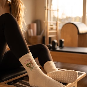 May include: Cream-colored crew socks featuring a ribbed texture and a graphic print of a green matcha bowl and a bamboo whisk. The text "MATCHA Girl" is printed in brown above and below the graphic. The soles have small white grip dots.
