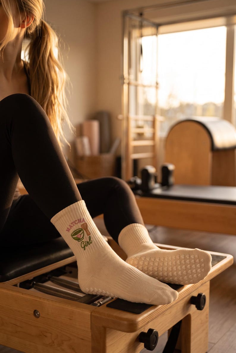 May include: Cream-colored crew socks featuring a ribbed texture and a graphic print of a green matcha bowl and a bamboo whisk. The text "MATCHA Girl" is printed in brown above and below the graphic. The soles have small white grip dots.