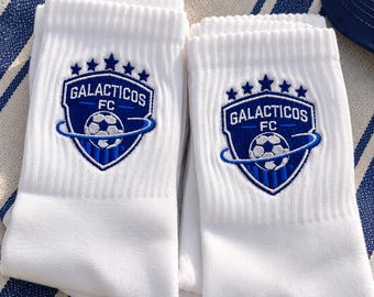 May include: A pair of white crew socks featuring an embroidered blue shield-shaped logo. The logo displays the text GALACTICOS FC, five stars across the top, and a central soccer ball graphic encircled by a ring, all rendered in blue and white thread.