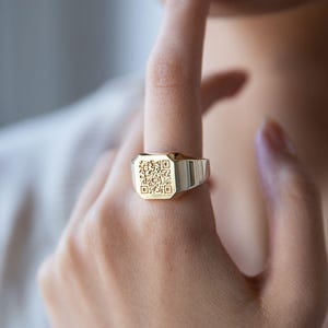 Könnte beinhalten: Ein goldener Siegelring mit einer quadratischen Fläche, die mit einem schwarz-weißen QR-Code verziert ist.