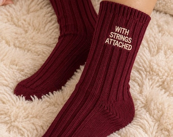 May include: A pair of deep burgundy ribbed knit socks featuring the text "WITH STRINGS ATTACHED" embroidered in white thread on the upper outer side of the ankle. The socks have a thick, textured knit construction and a mid-calf length design.