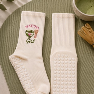 May include: A pair of cream-coloured ribbed crew socks featuring an embroidered graphic of a green matcha bowl with a pink heart and a bamboo whisk. The text "MATCHA Girl" is embroidered above and below the graphic. The soles have heart-shaped grips.