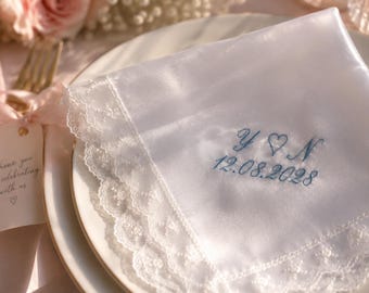 May include: A white square handkerchief with a scalloped lace border, featuring blue embroidered initials Y and N separated by a heart, with the date 12.08.2028 embroidered below in a cursive script, resting on a white ceramic plate.