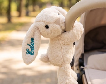 May include: A cream-colored plush rabbit toy with long ears, black eyes, and a small nose. The name Eden is embroidered in teal cursive script on one of the rabbit's ears. The stuffed animal is hanging onto the beige handle of a baby stroller.