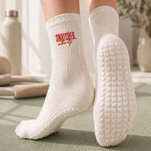 May include: A pair of cream-coloured ribbed crew socks featuring the red and gold embroidered text "SNATCHED in the city" on the outer ankle. The soles of the socks are covered in a repeating pattern of small, raised white heart-shaped silicone grips.