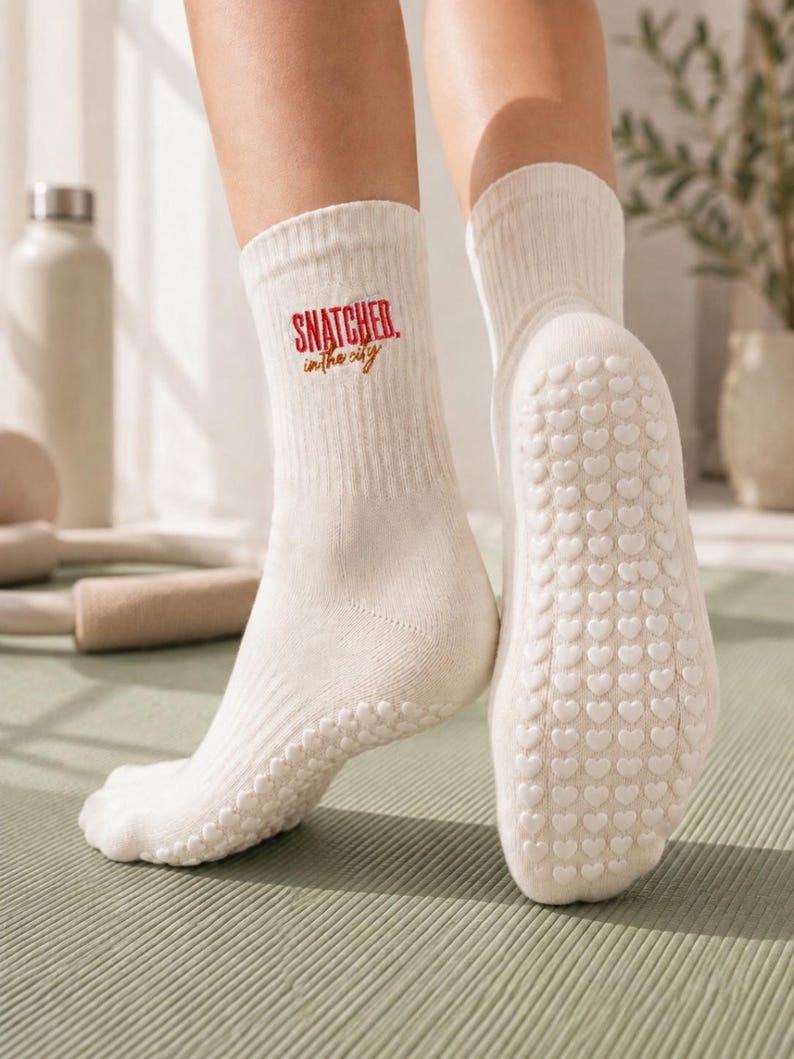 May include: A pair of cream-coloured ribbed crew socks featuring the red and gold embroidered text "SNATCHED in the city" on the outer ankle. The soles of the socks are covered in a repeating pattern of small, raised white heart-shaped silicone grips.