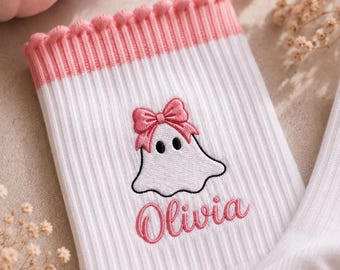 May include: White ribbed crew socks featuring a pink pom-pom trim at the cuff. The socks are embroidered with a white ghost character wearing a pink bow and the name Olivia in pink script lettering. The sole features small pink heart-shaped grips.