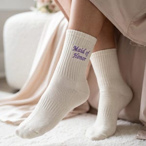 May include: A pair of cream-colored ribbed crew socks featuring the text "Maid of Honor" embroidered in purple thread on the outer side of the ankle. The socks have a classic knit texture and reach mid-calf, providing a simple and clean appearance.