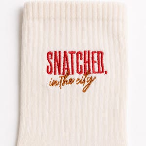 May include: A cream-coloured ribbed sock featuring embroidered text. The word SNATCHED is stitched in bold red capital letters, while the phrase in the city is stitched below in a smaller, cursive brown font, centred on the fabric.