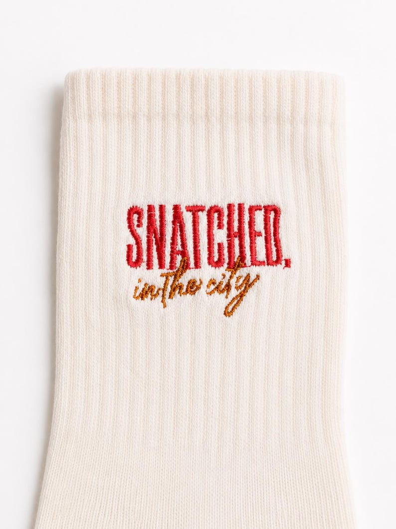May include: A cream-coloured ribbed sock featuring embroidered text. The word SNATCHED is stitched in bold red capital letters, while the phrase in the city is stitched below in a smaller, cursive brown font, centred on the fabric.
