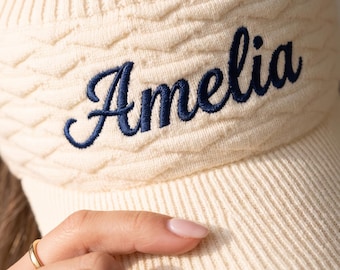 May include: A cream-colored knit visor featuring a textured cable knit pattern on the headband and a ribbed brim. The name Amelia is embroidered in dark blue cursive script across the front center of the headband, providing a clear contrast.