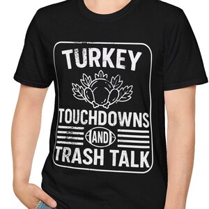 Op de afbeelding: Zwart T-shirt met een witte opdruk met de tekst "TURKEY TOUCHDOWNS AND TRASH TALK". De afbeelding bevat een illustratie van een kalkoen. De persoon op de foto draagt een blauwe spijkerbroek.