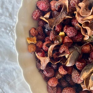 May include: A white scalloped bowl filled with a collection of dried rose hips and dark brown curled botanical elements. The dried fruit pieces are textured, round, and vary in shades of deep red, burgundy, and brown, creating a dense, rustic display.