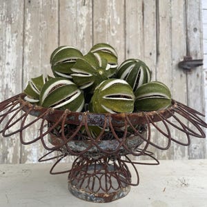 May include: A rustic wire basket filled with dried green citrus fruit. The basket has a decorative scalloped edge and a pedestal base.