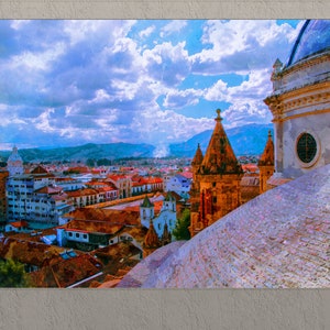 May include: A panoramic view of a city from the top of a building with a tiled roof. The city is filled with colorful buildings and a mountain range in the background. The sky is blue with white clouds.