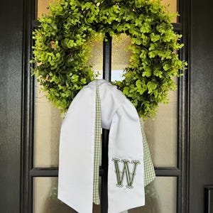May include: A green leafy wreath hangs on a black door. A white sash with a green and white gingham trim is draped over the wreath. The sash has a large embroidered green letter "W".