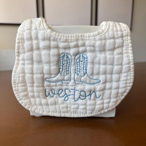 Puede incluir: Babero de bebé acolchado blanco con botas de vaquero bordadas en azul y el nombre "Weston". El babero tiene forma redondeada y una textura suave, ideal para bebés.